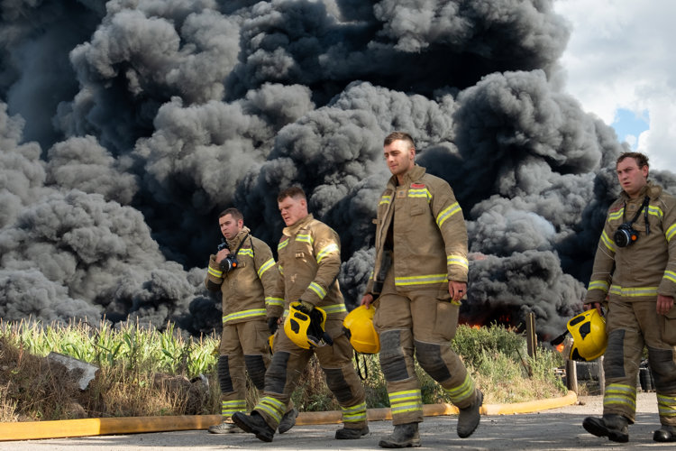 Four firefighters walking down a road with a large fire in background