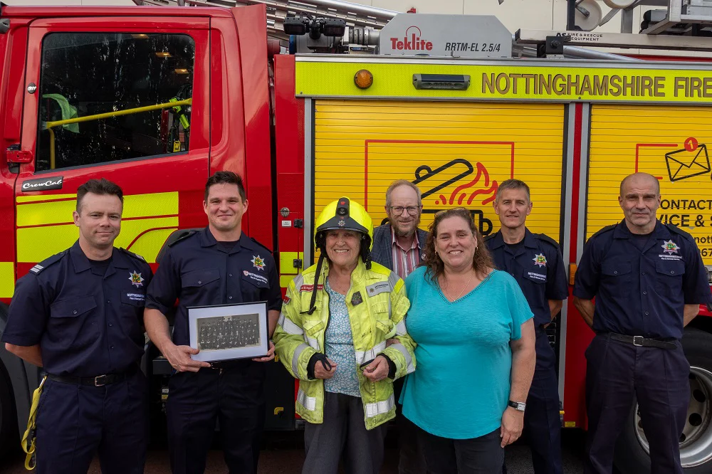 firefighters and three members of the public stand beside a fire engine smiling at the camera