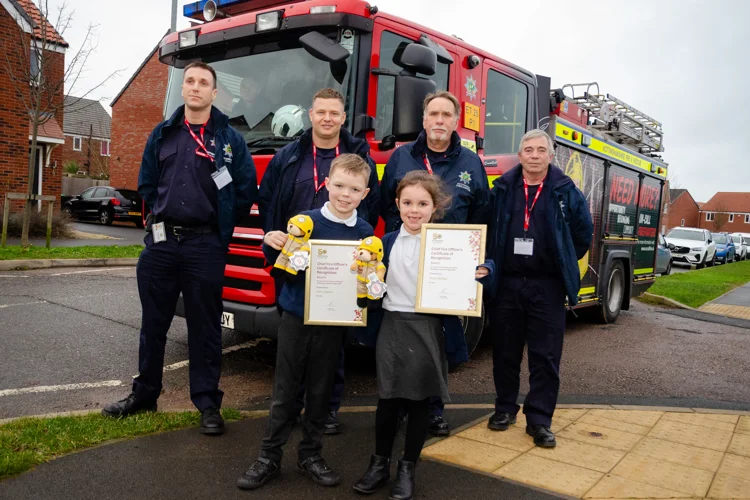 Photo of four firefighters and two children holding certificates in front of a fire engine.