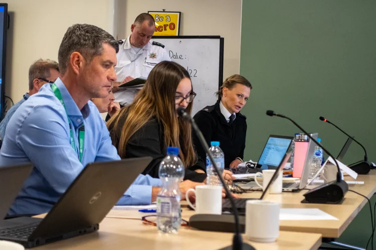 A police officer speaks into a microphone with others listening and a fire officer taking notes.