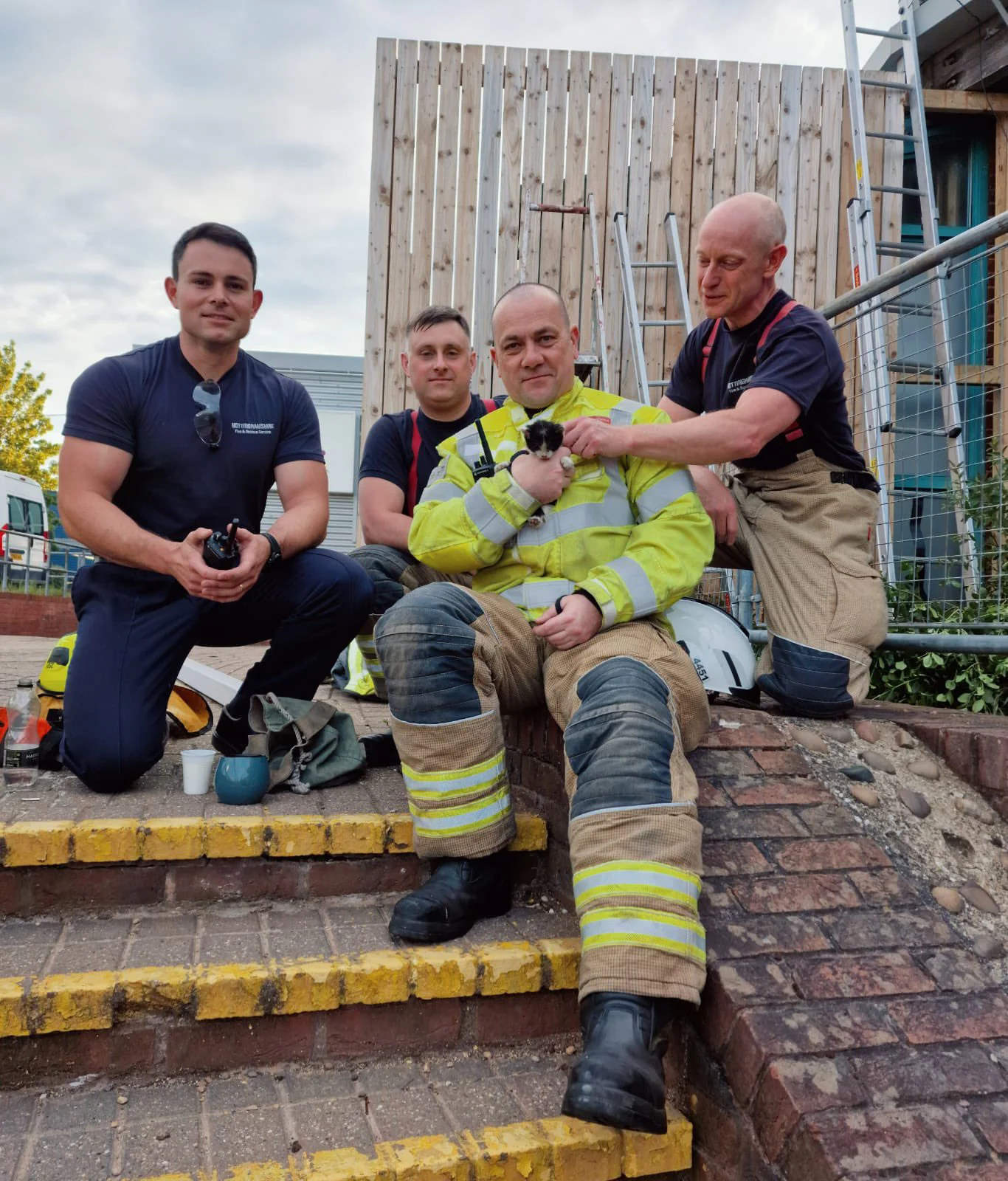 four firefighters sit on steps, the one in the middle holds a tiny black and white kitten, while other leans over to stroke the kitten