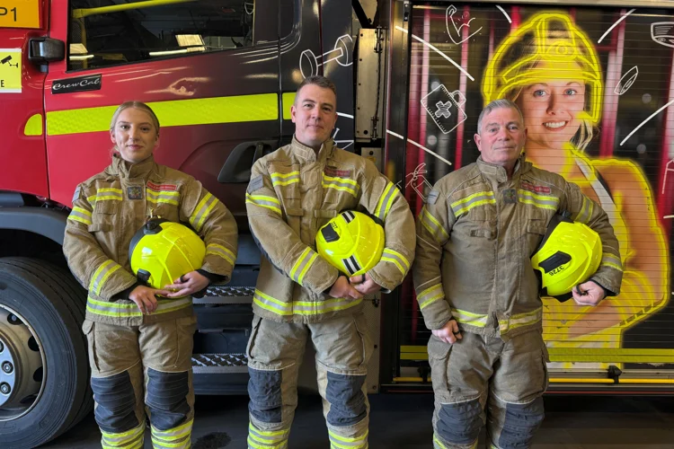 Three firefighters stood at the side of the fire engine in fire kit holding their helmets