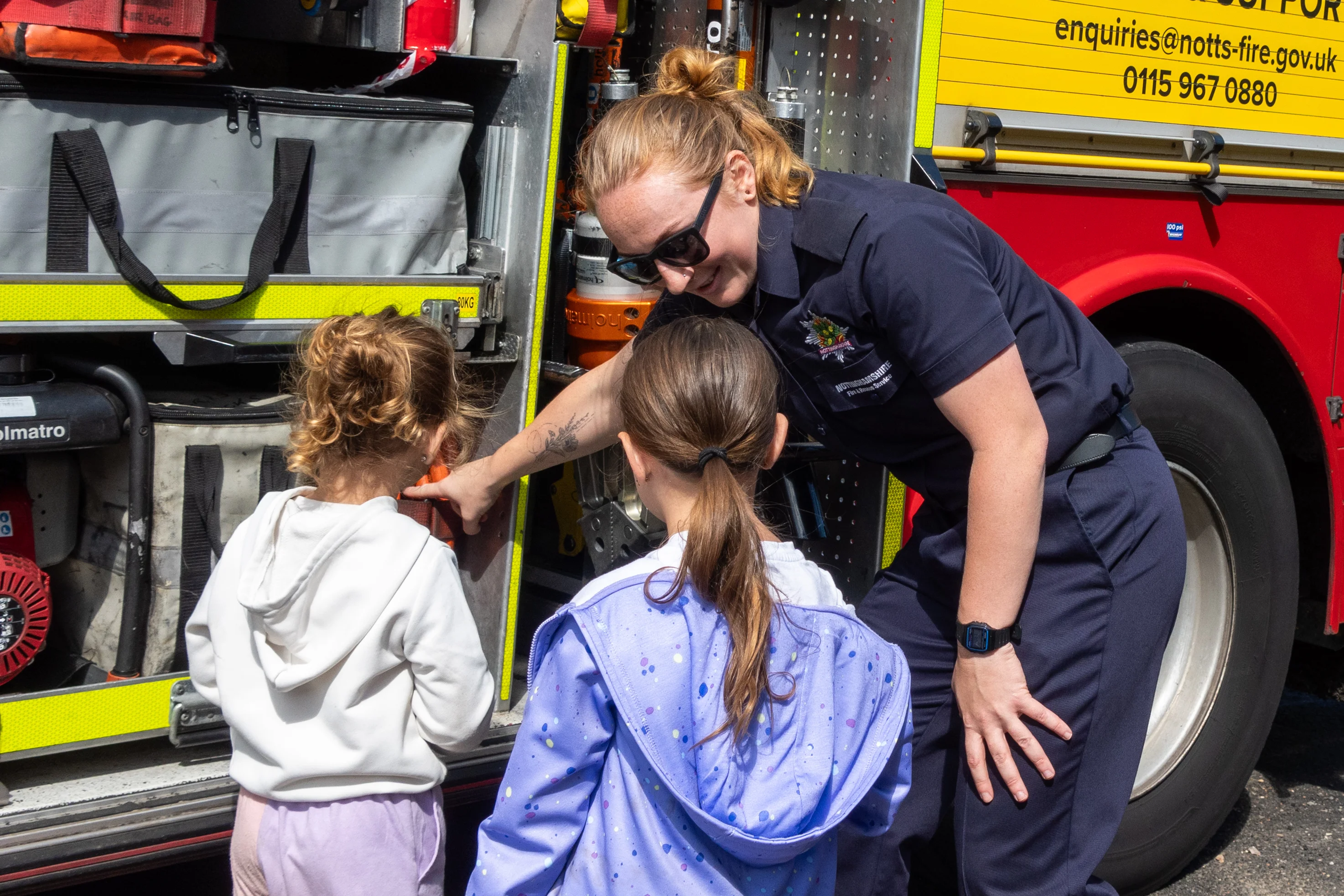 A firefighter shows two young girls one of the lockers on the fire engine.
