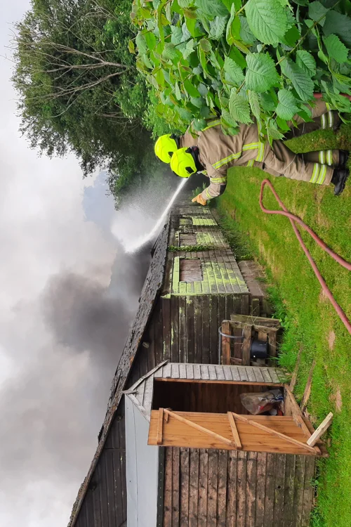 two firefighters point towards a barn on fire, spraying a hose towards the smoke