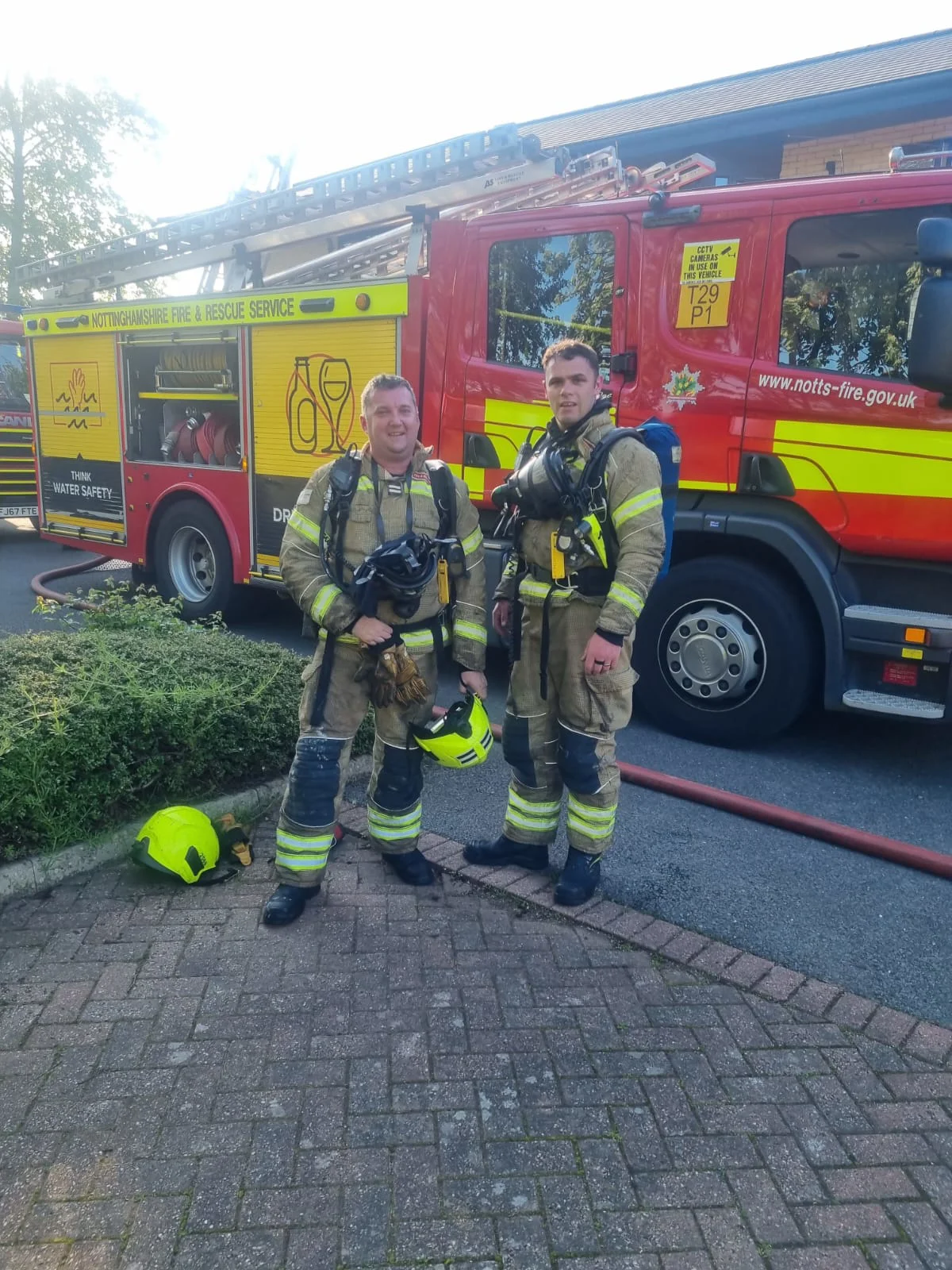 two firefighters stand next to a fire engine wearing full fire kit