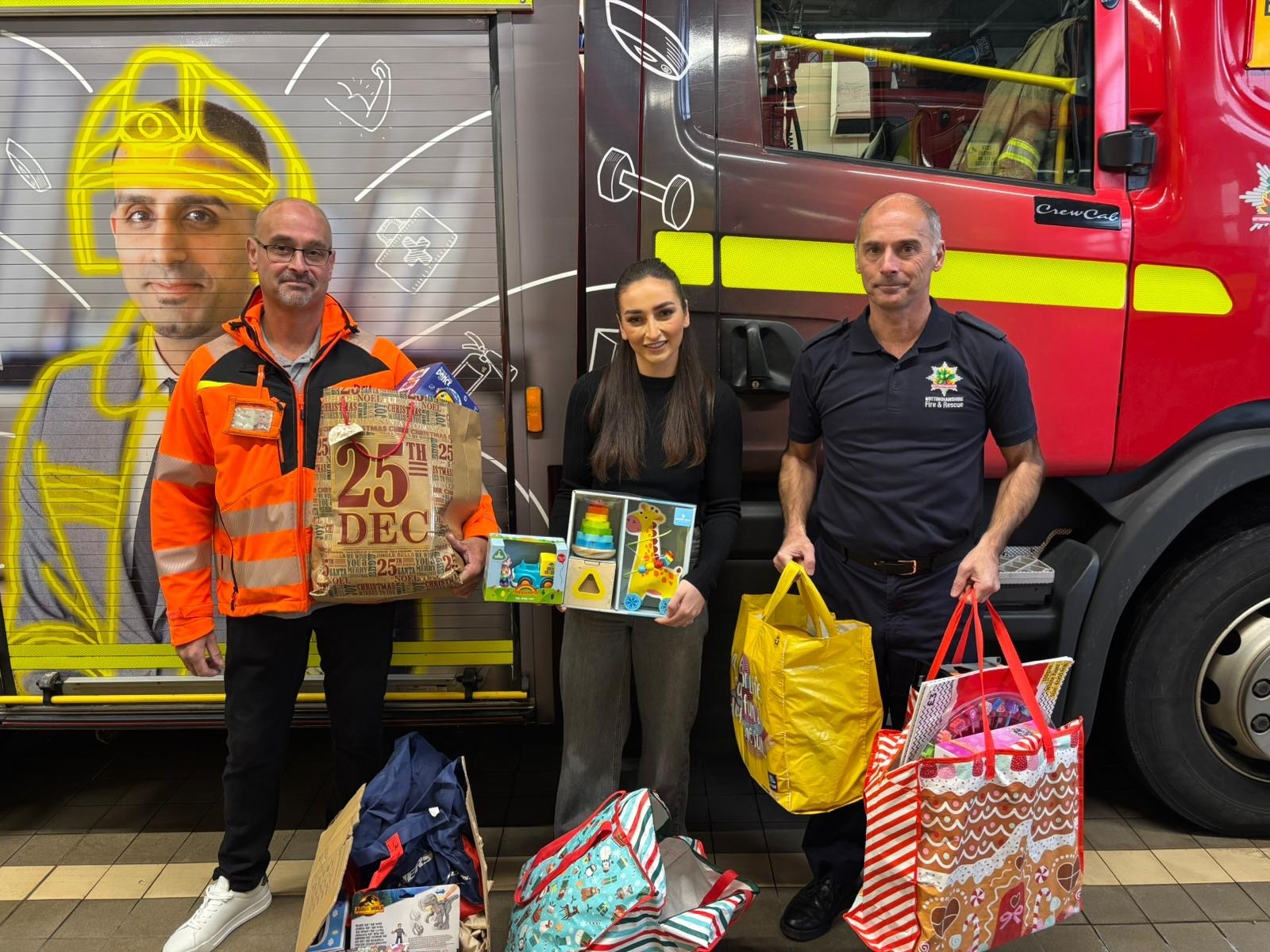A man in a high-vis, a female, and a firefighter holding toys in front of the fire engine.