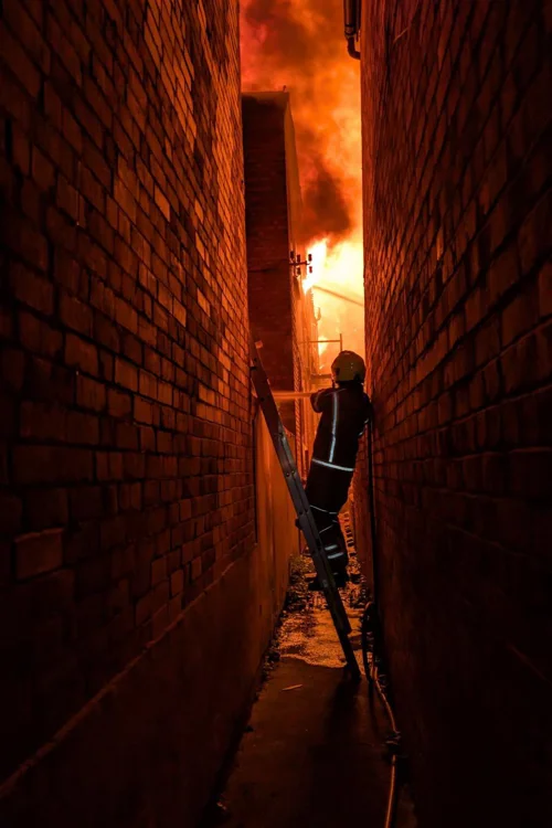 Orange photograph of firefighter on a ladder with fire in the background.