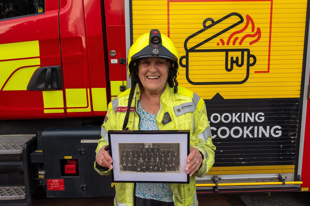 a lady smiles looking at the camera while holding a picture of a group of firefighters, and wearing fire kit