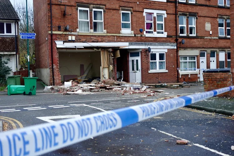 Police tape in the foreground looking to a front row of houses with one ground floor blown out with rubble and debris on the floor.