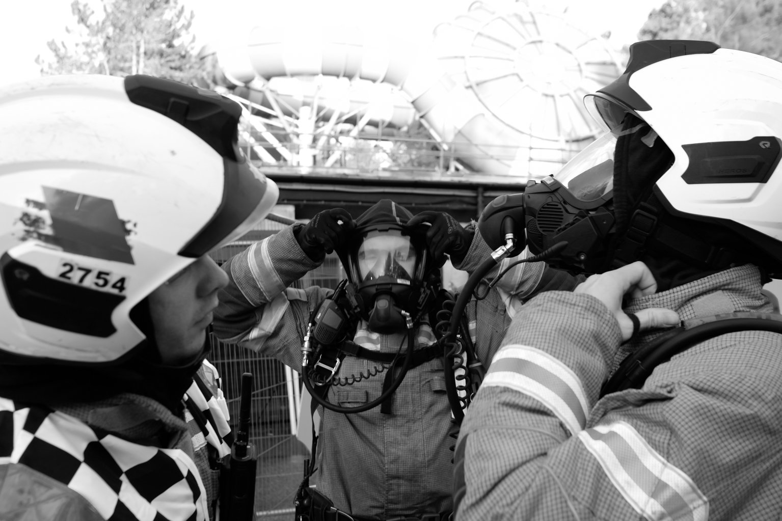 Black and white image of two firefighters putting on breathing apparatus whilst being inspected by the entry control officer.
