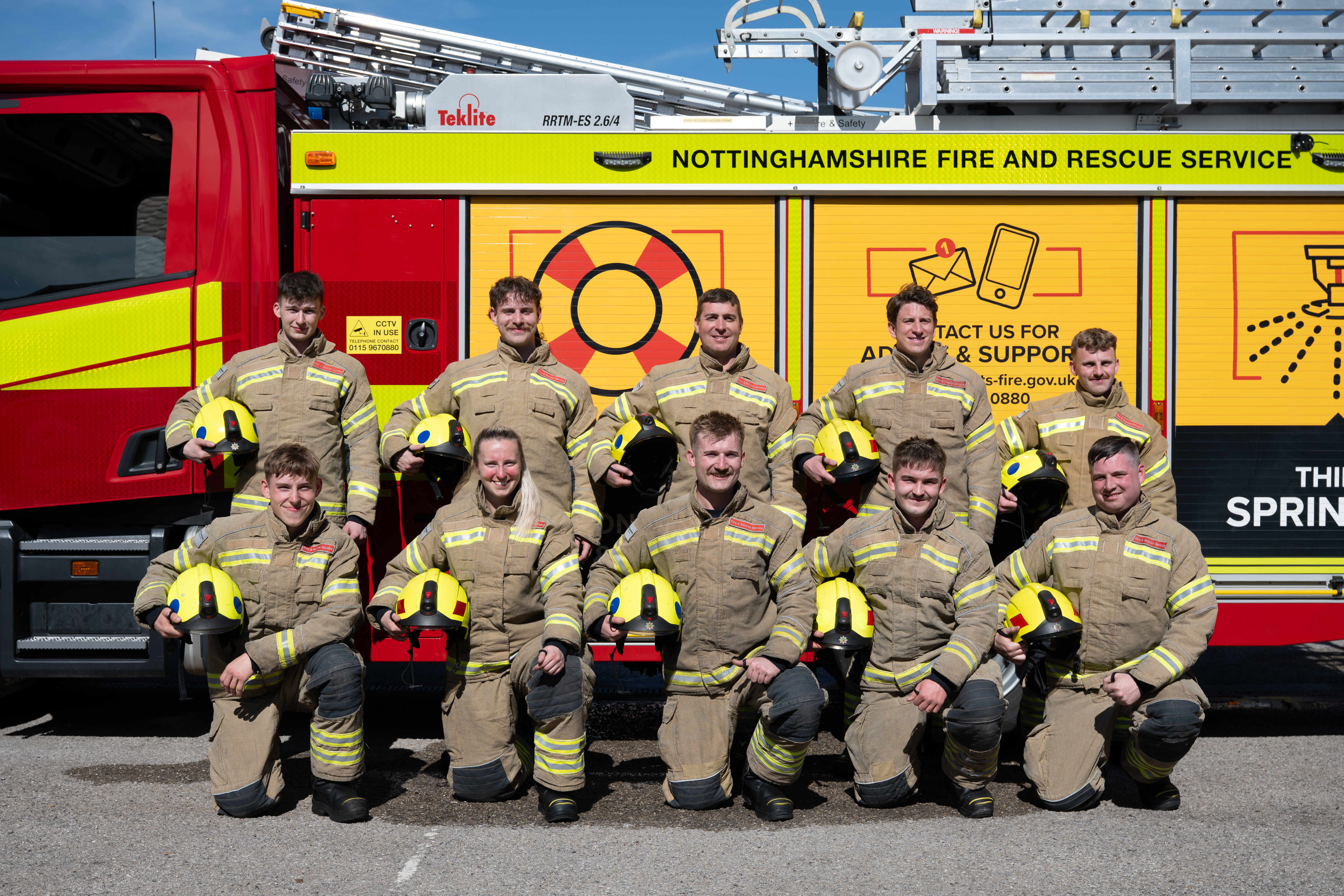 10 firefighters in fire kit infront of the engine in two rows holding their helmets