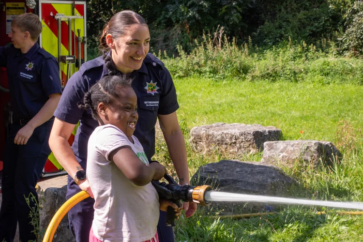 A firefighter and young girl smile as the girl squirts water from the hose.