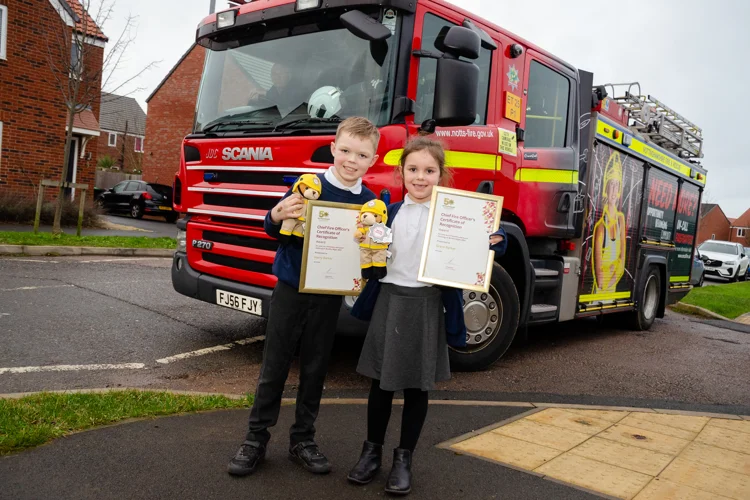 A young boy and girl in front of a fire engine smiling with certificates.