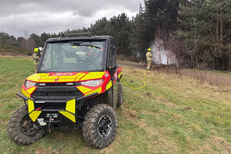Landscape photo of fire service red and yellow polaris in the foreground, with firefighter putting out fire in the background.