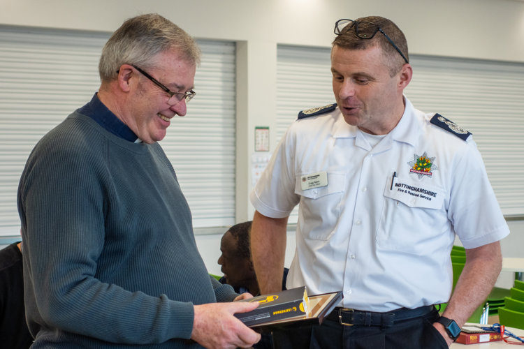 David Milner holds his certificate and other gifts, smiling, as Chief Fire Officer Craig Parkin speaks to him.